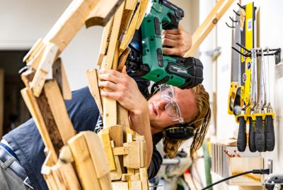 A student wearing safety goggles and ear protection uses a nail gun to assemble a wooden animatronic sculpture in a workshop, surrounded by hand tools hanging on the wall.